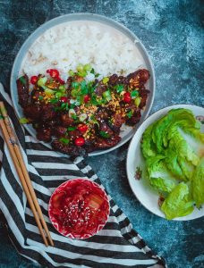 A delicious plate of pork bulgogi topped with green onions and chili with white rice, accompanied by a bowl of fresh lettuce, chopsticks, and a small bowl of red kimichi on a striped cloth.