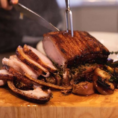 A person is slicing a cooked piece of Porchetta on a wooden cutting board with vegetables.