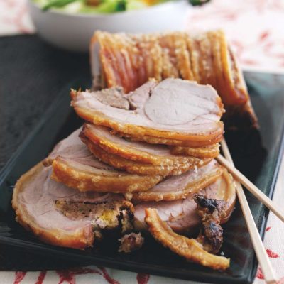 A succulent roast loin of pork with oriental stuffing, sliced and beautifully arranged on a black plate, accompanied by chopsticks. In the background, a bowl of vibrant, fresh vegetables.
