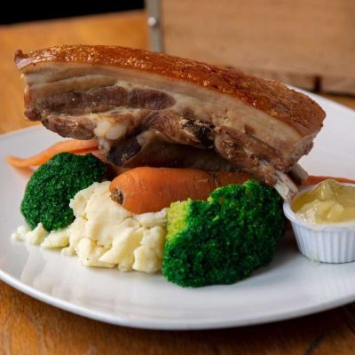 A succulent piece of super slow roasted pork belly with crispy crackling, served alongside vibrant green broccoli, tender carrots, creamy mashed potatoes, and a side of rich sauce.