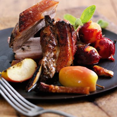 A succulent grilled pork chop accompanied by roasted apples and colorful bell peppers, garnished with fresh green herbs, served on a dark plate.