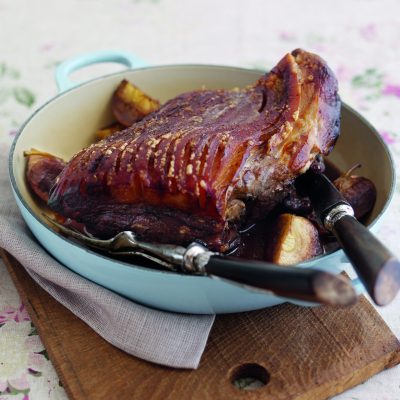 A succulent slow-roasted pork shoulder with a rich glaze, accompanied by roasted apples and seasoned with ginger, treacle, and allspice, served in a blue pan on a wooden board over a floral tablecloth.