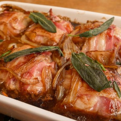 A baked dish of pork faggots and liver balls wrapped in bacon and garnished with sage leaves in a white baking dish.