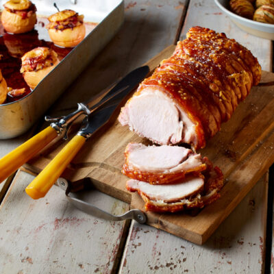A sliced roast pork loin on a wooden cutting board, with a carving fork and knife, next to a baking tray of baked apples and a bowl of hasselback potatoes on a rustic table.