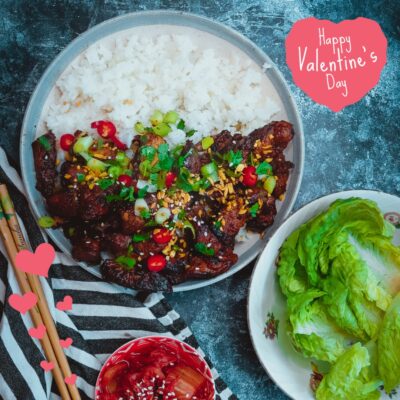 A delicious plate of pork bulgogi garnished with fresh herbs and vegetables, served next to a bowl of lettuce and a dish of sauce, with a Valentine’s Day greeting in the background.