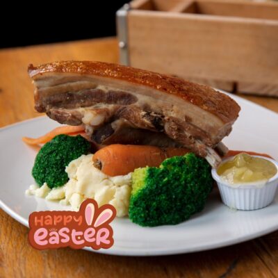 A festive Easter meal featuring a pork belly with crispy crackling, creamy mashed potatoes, cooked carrots and broccoli, served on a white plate. The dish is accompanied by a small cup of apple sauce, and a “Happy Easter” greeting is displayed at the bottom of the image.
