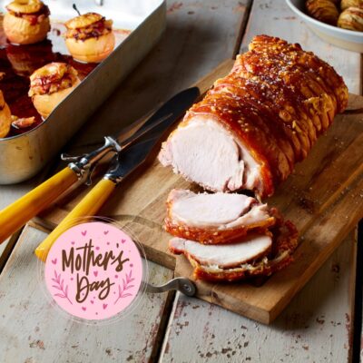 A succulent roasted pork loin, sliced and resting on a wooden cutting board, with a carving fork and knife. In the background, there are baked goods in a tray. A pink tag with “Mother’s Day” written on it is prominently displayed in the foreground.